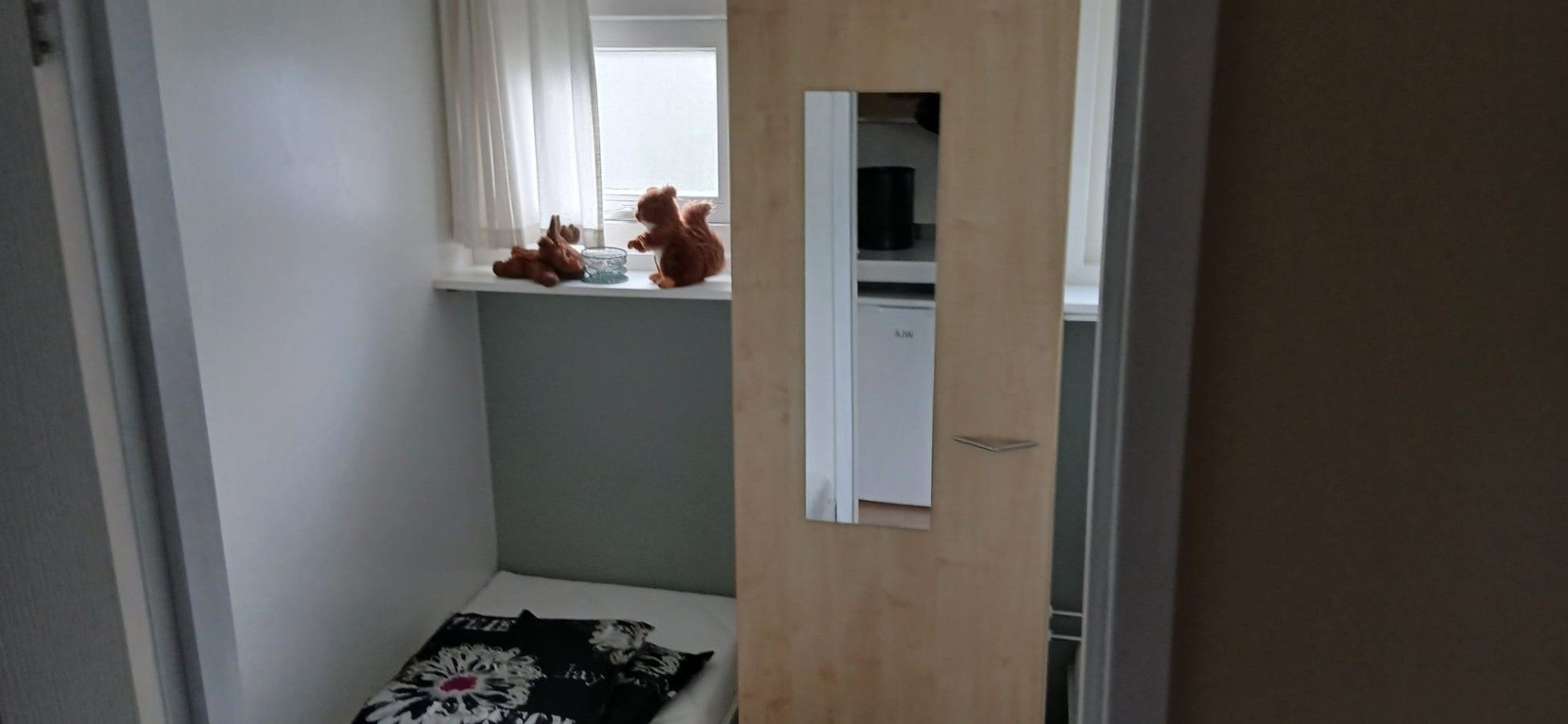 A narrow room with a window, a small bed with a patterned blanket, and a teddy bear on the windowsill.