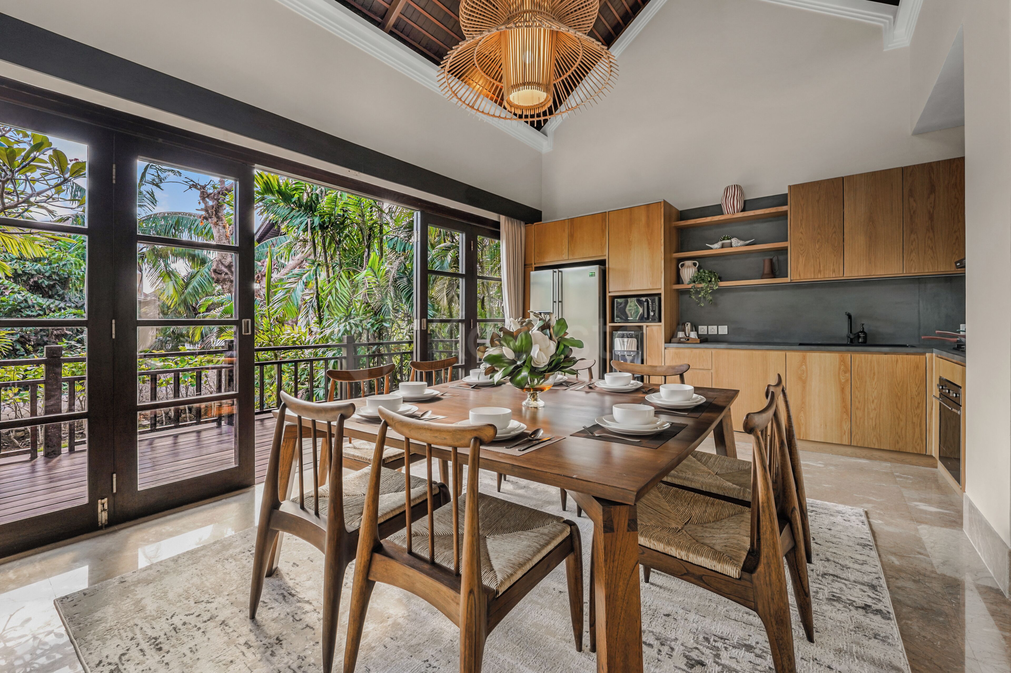 Una cocina moderna con armarios de madera, un comedor con una mesa grande y sillas, y vistas a la vegetación exuberante a través de puertas correderas de vidrio.