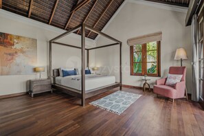 A four-poster bed with a wooden frame, a small bedside table with a lamp, a pink armchair, and a window with a view of greenery.