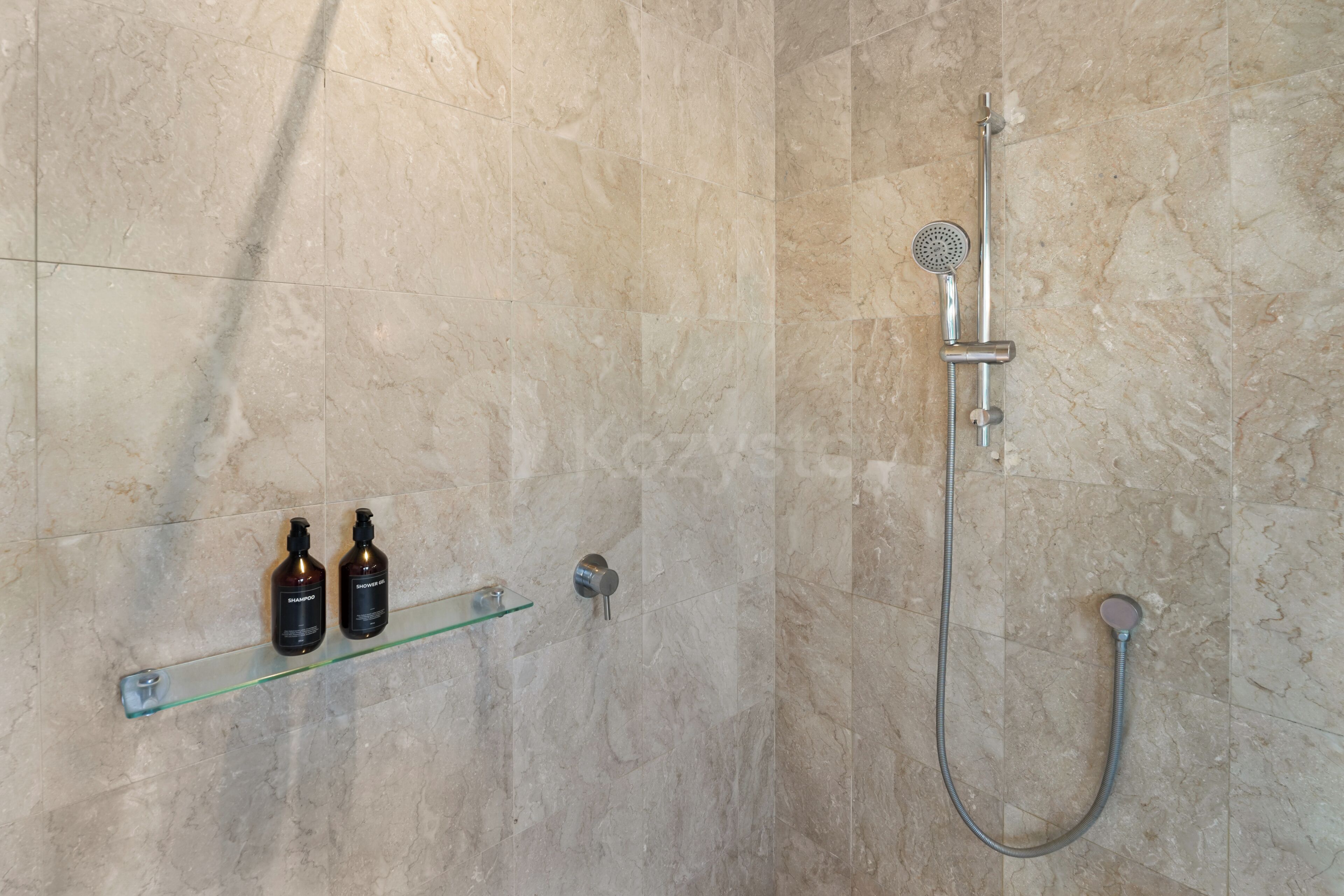 A shower area with a handheld showerhead, a wall-mounted showerhead, and a glass shelf with two bottles.
