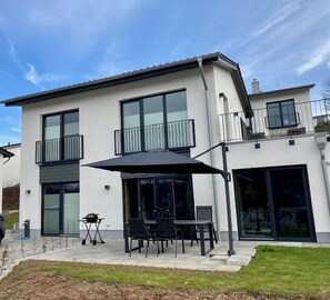 A modern two-story house with a patio area featuring a table and chairs, a grill, and a large umbrella. - SEEside13. Lake view. Sauna. Front Row at Diemelsee (Diemelsee)