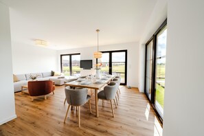 Un salon moderne doté d’une grande fenêtre, d’un coin repas avec une table et des chaises en bois, et d’un canapé gris.