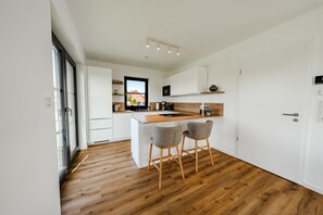 A modern kitchen with white cabinetry, a wooden countertop, and two bar stools. - Hildes Huuske | Right on the Water - Outdoor Sauna (Greetsiel)