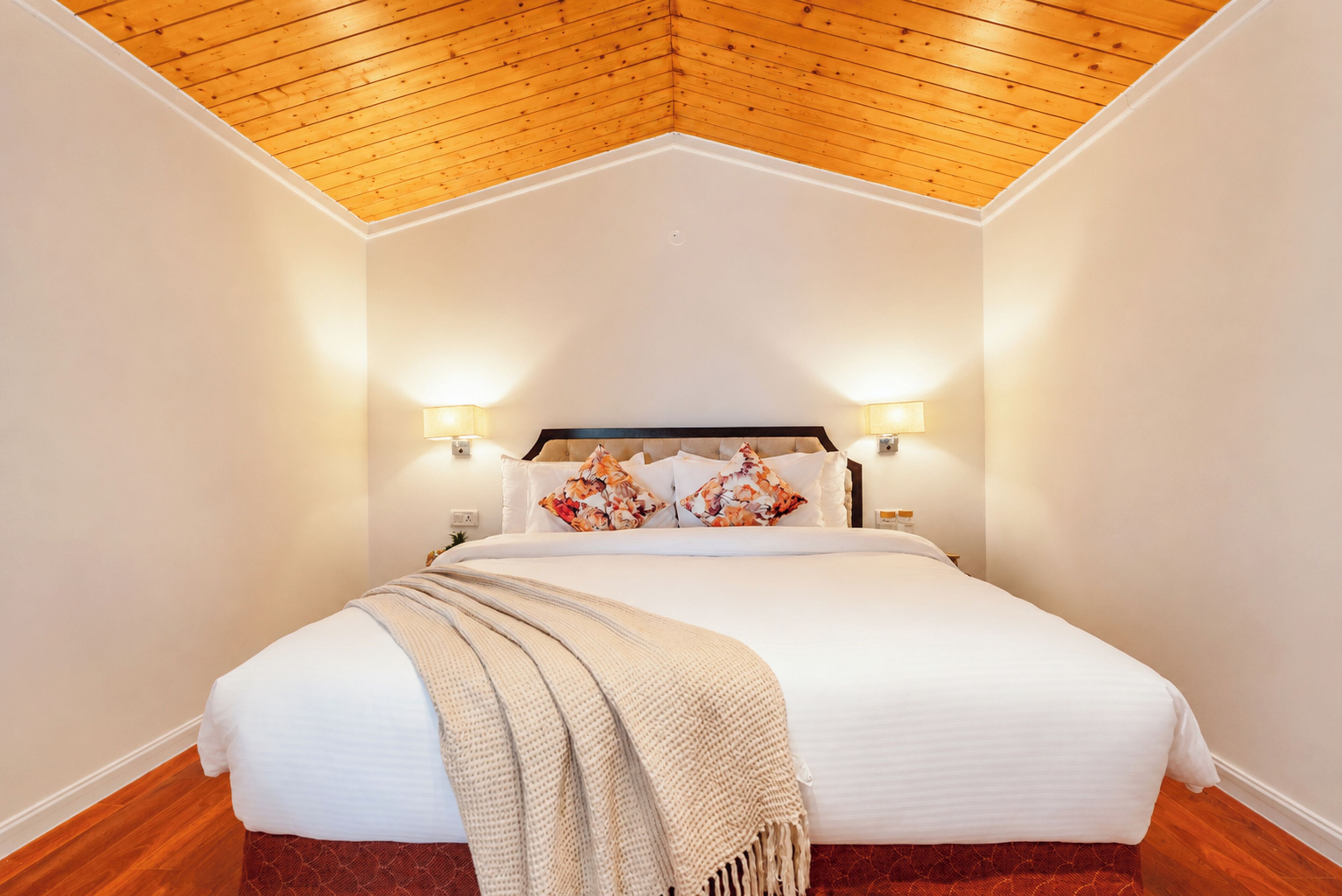 Habitación de hotel con techo de madera, cama con ropa blanca y almohadas estampadas, y lámparas fijadas a la pared.