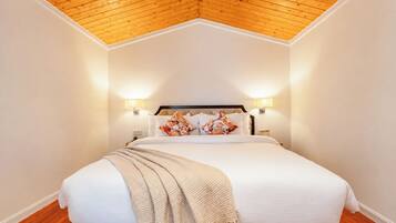 Habitación de hotel con techo de madera, cama con ropa blanca y almohadas estampadas, y lámparas fijadas a la pared.