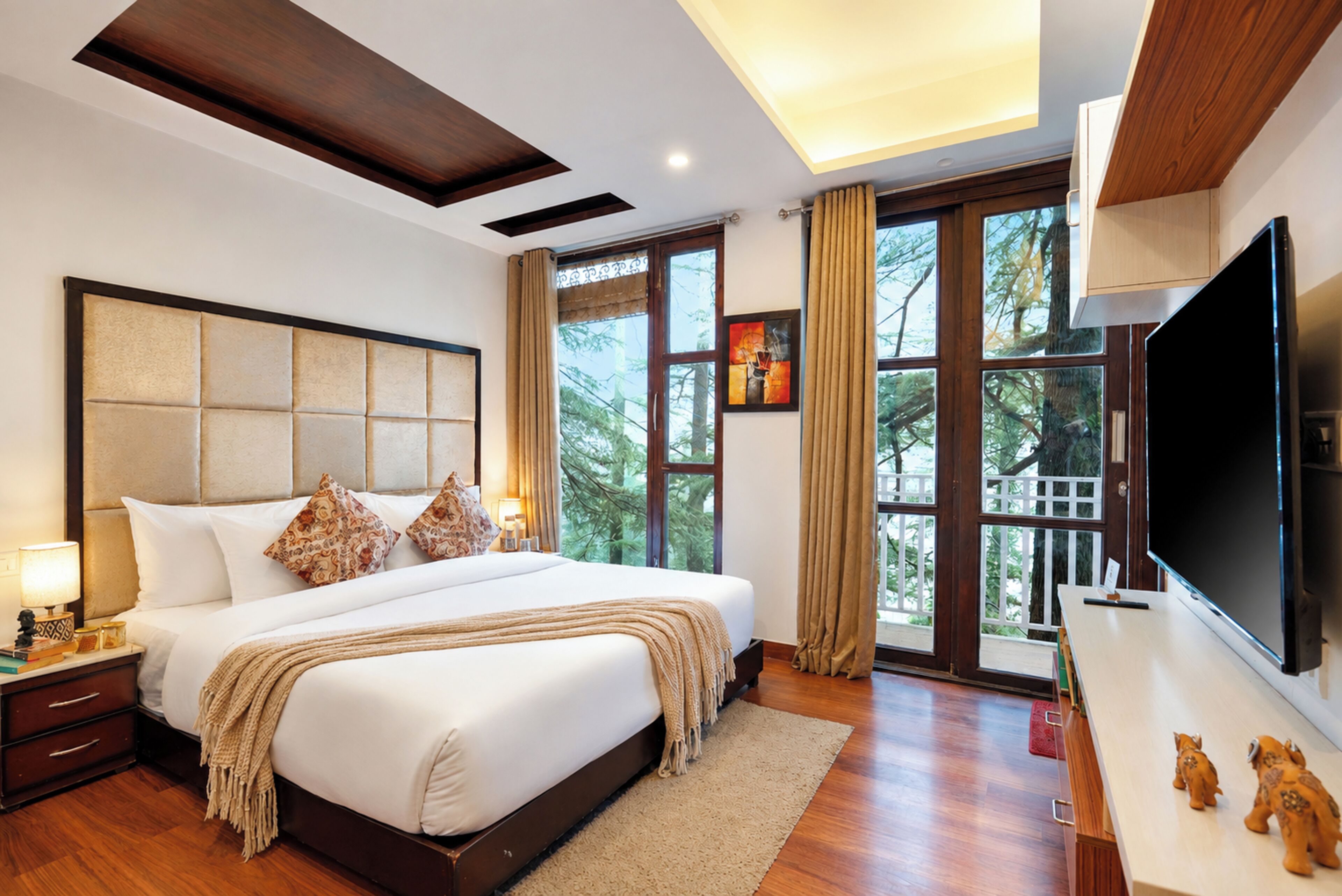 A hotel room with a large bed, a flat-screen TV, a nightstand, and a window with a view of trees.