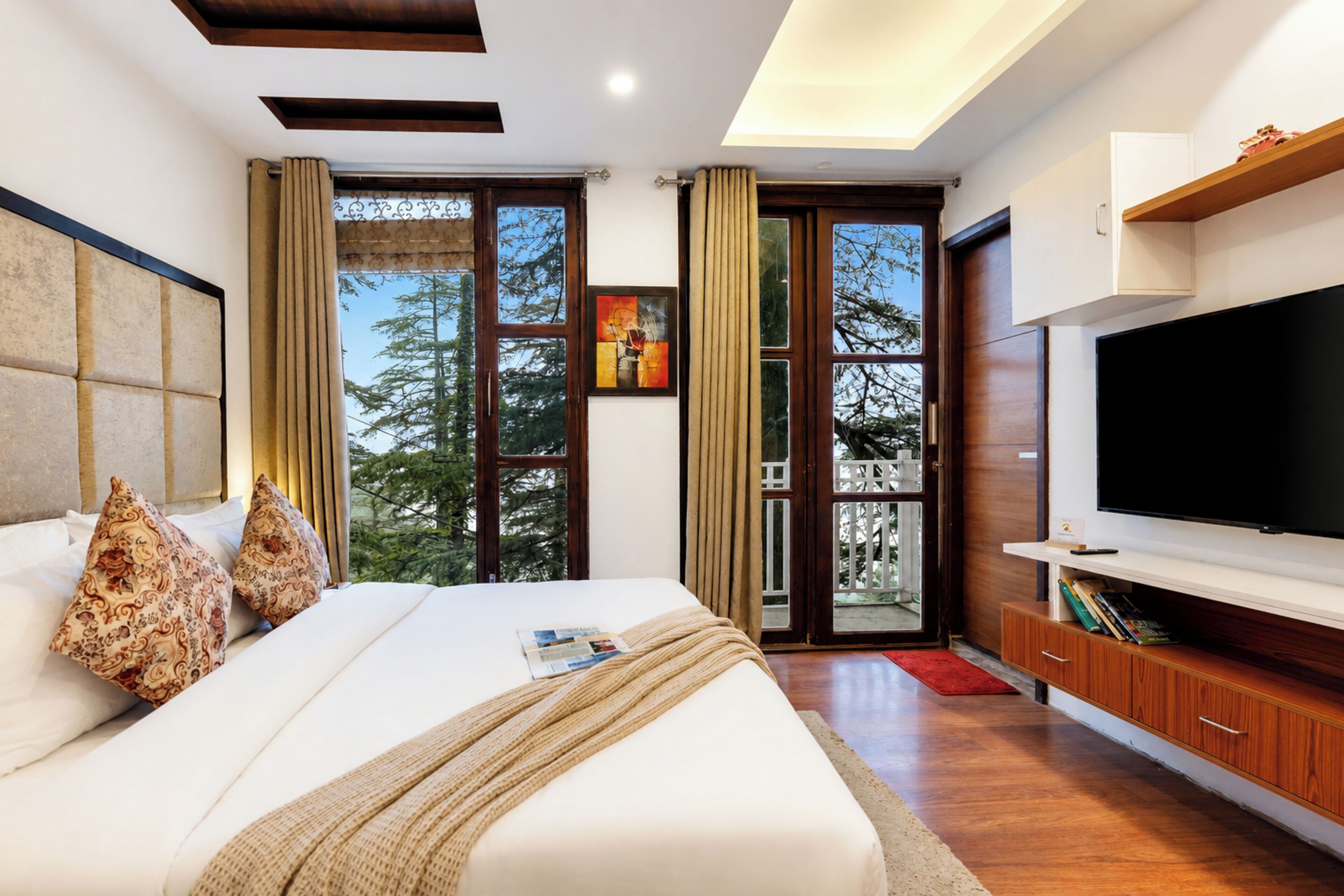 A modern bedroom with a large bed, a flat-screen TV, and a view of trees through the windows.