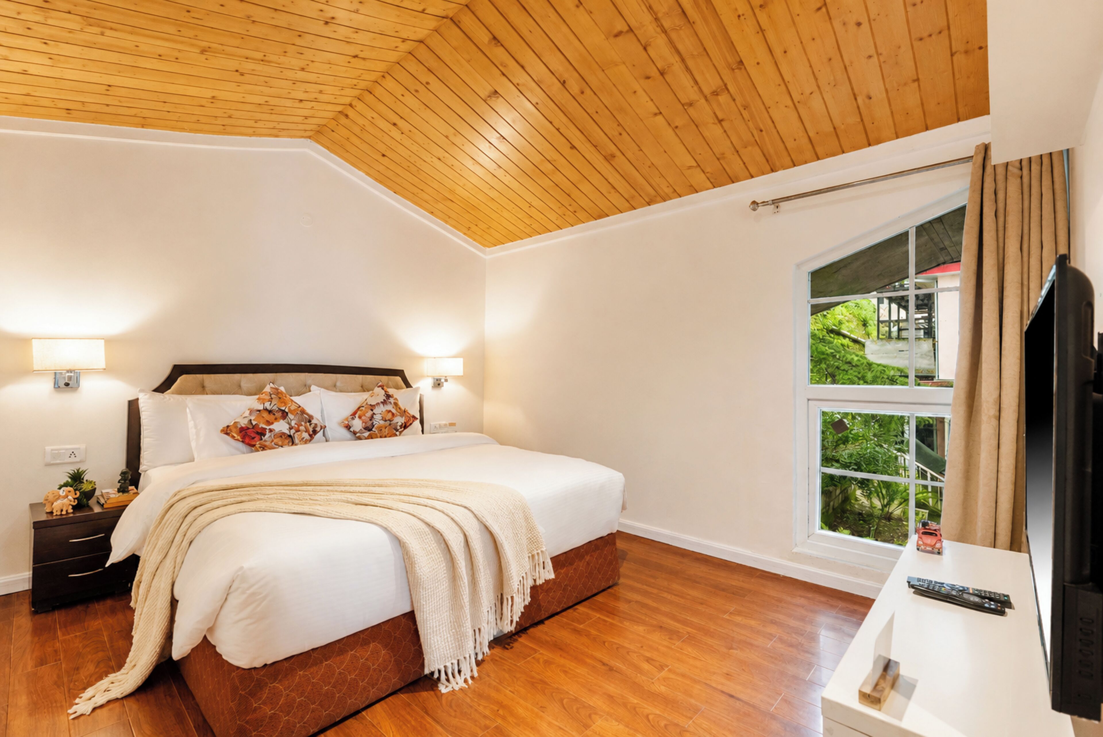 Un dormitorio con techo de madera, una cama grande con ropa blanca, dos lámparas de noche, una mesita de noche con lámpara, una ventana con cortinas y un mueble para televisión con control remoto.