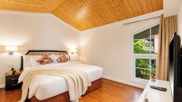 Un dormitorio con techo de madera, una cama grande con ropa blanca, dos lámparas de noche, una mesita de noche con lámpara, una ventana con cortinas y un mueble para televisión con control remoto.