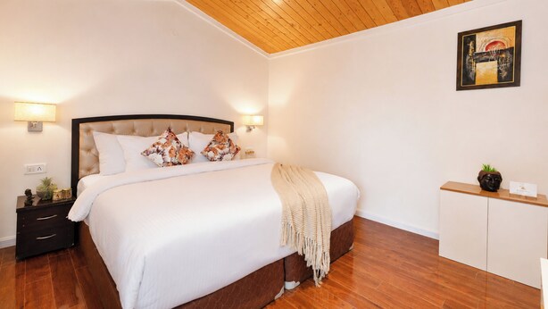 A bedroom with a wooden ceiling, a bed with white bedding, two bedside lamps, a nightstand with a lamp, a small cabinet, and a framed picture on the wall.