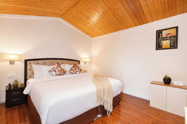 A bedroom with a wooden ceiling, a bed with white bedding, two bedside lamps, a nightstand with a lamp, a small cabinet, and a framed picture on the wall.