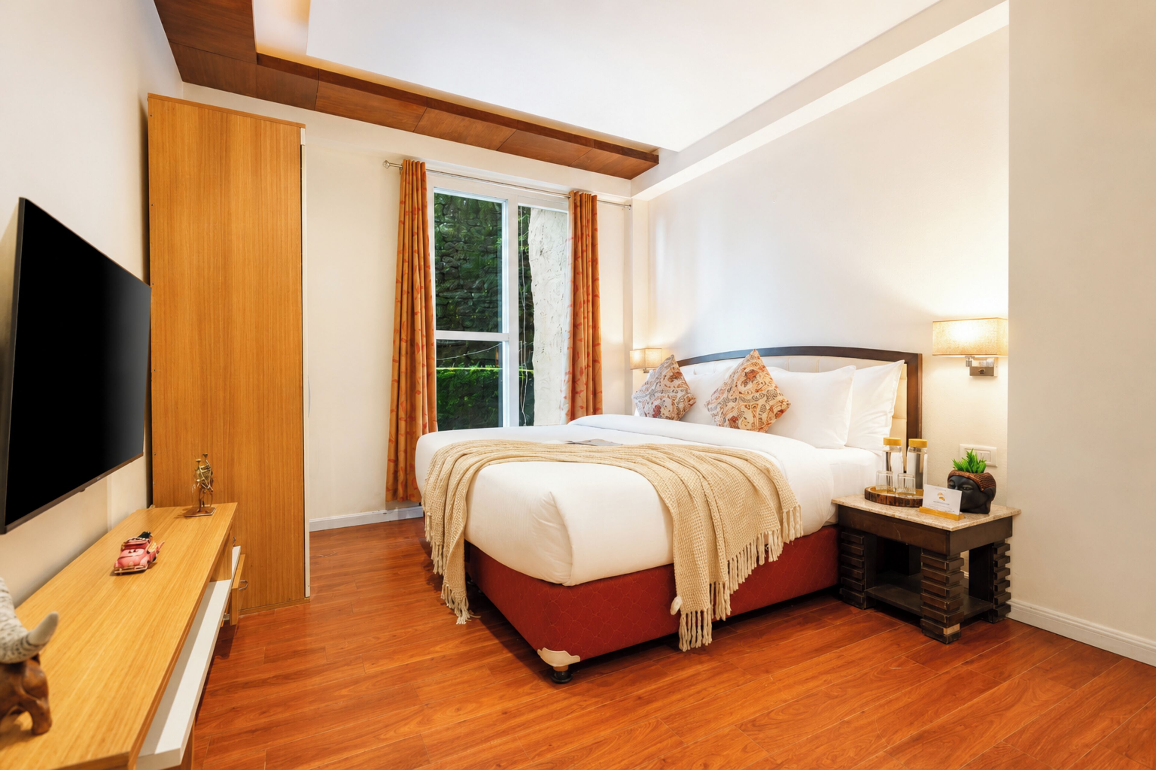 A hotel room with a large bed, a TV, a bedside table with a lamp, and a window with curtains.