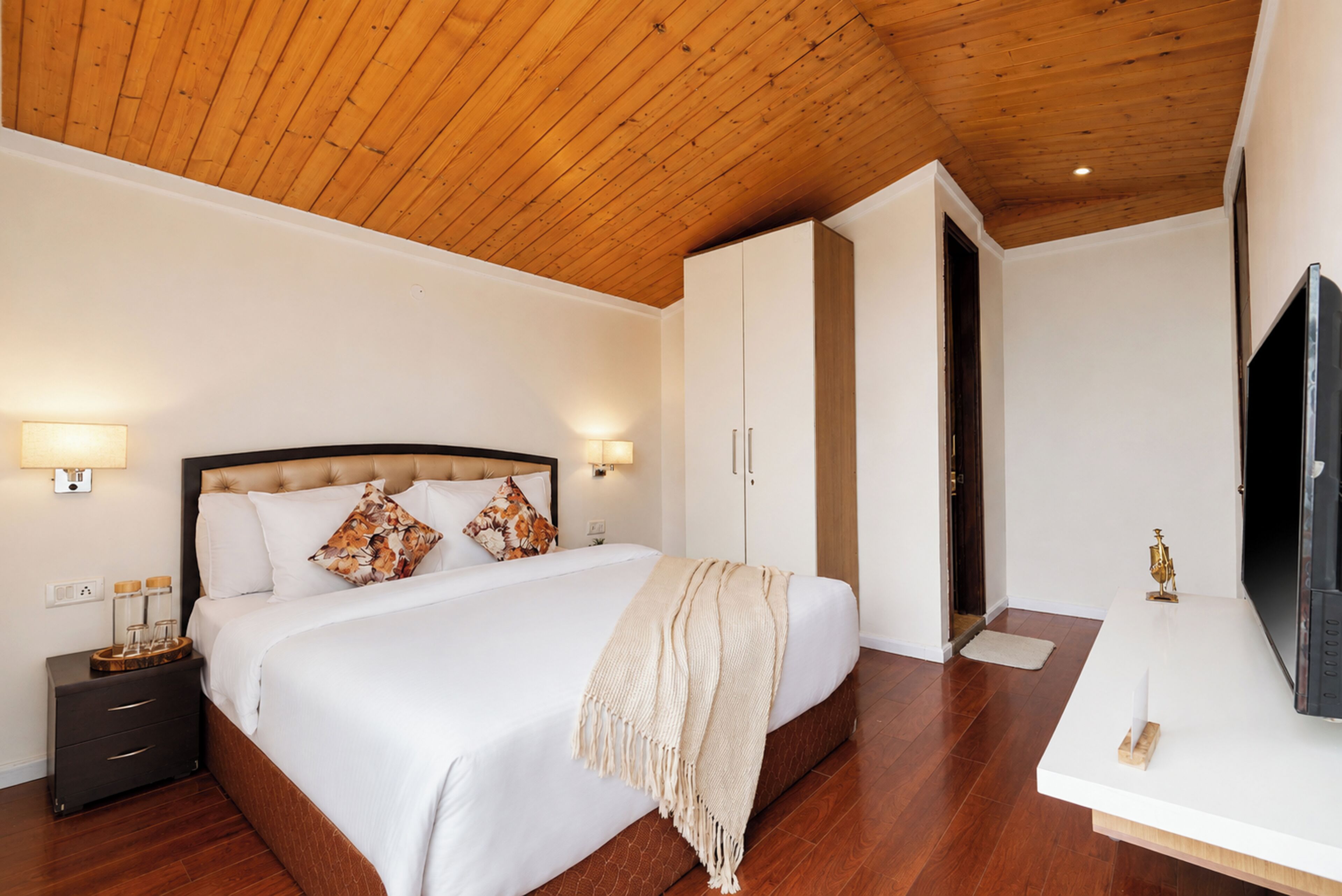 Un dormitorio con techo de madera, cama con ropa de cama blanca, mesita de noche, armario y televisor.