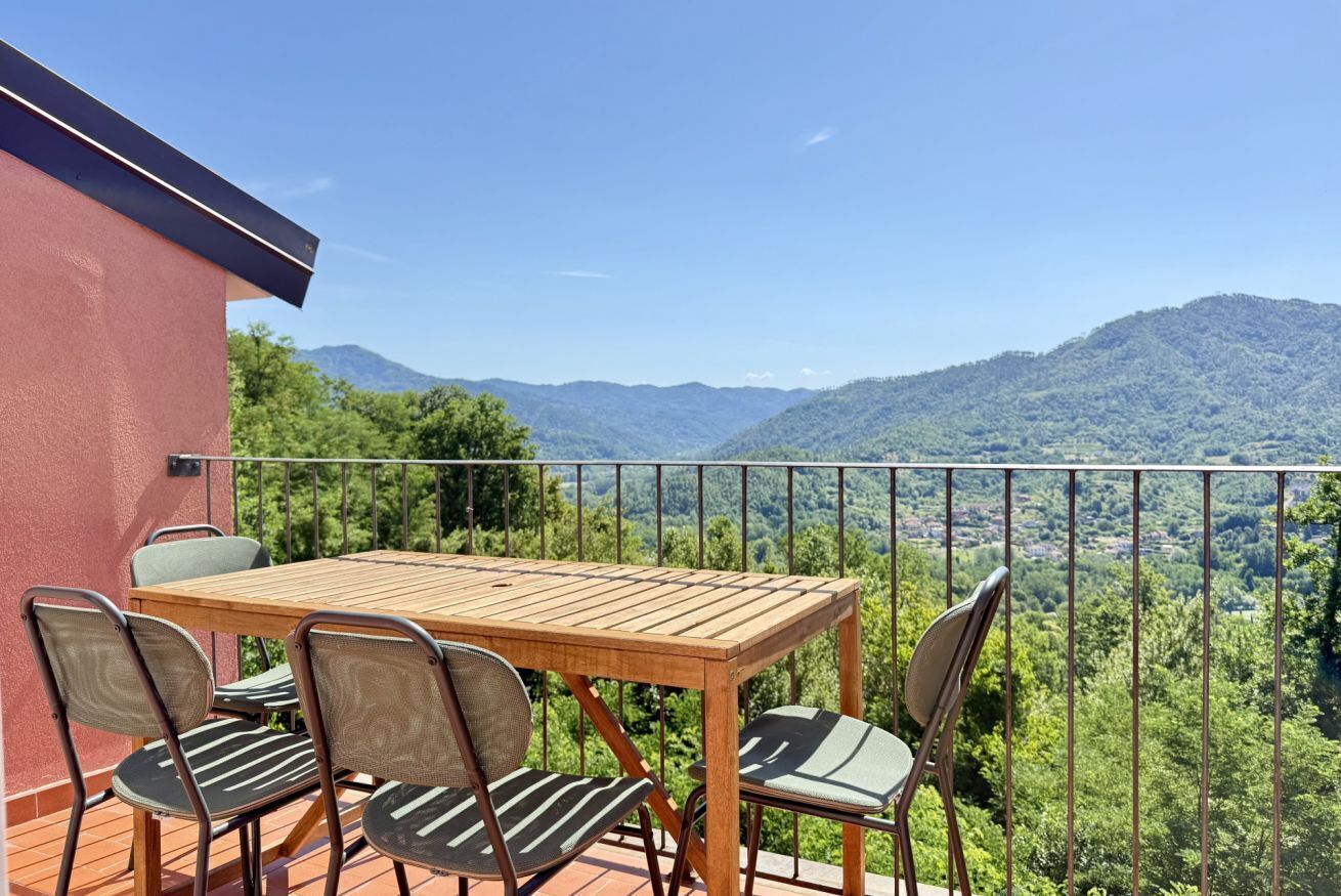 A wooden table and chairs on a balcony with a view of green hills and mountains.