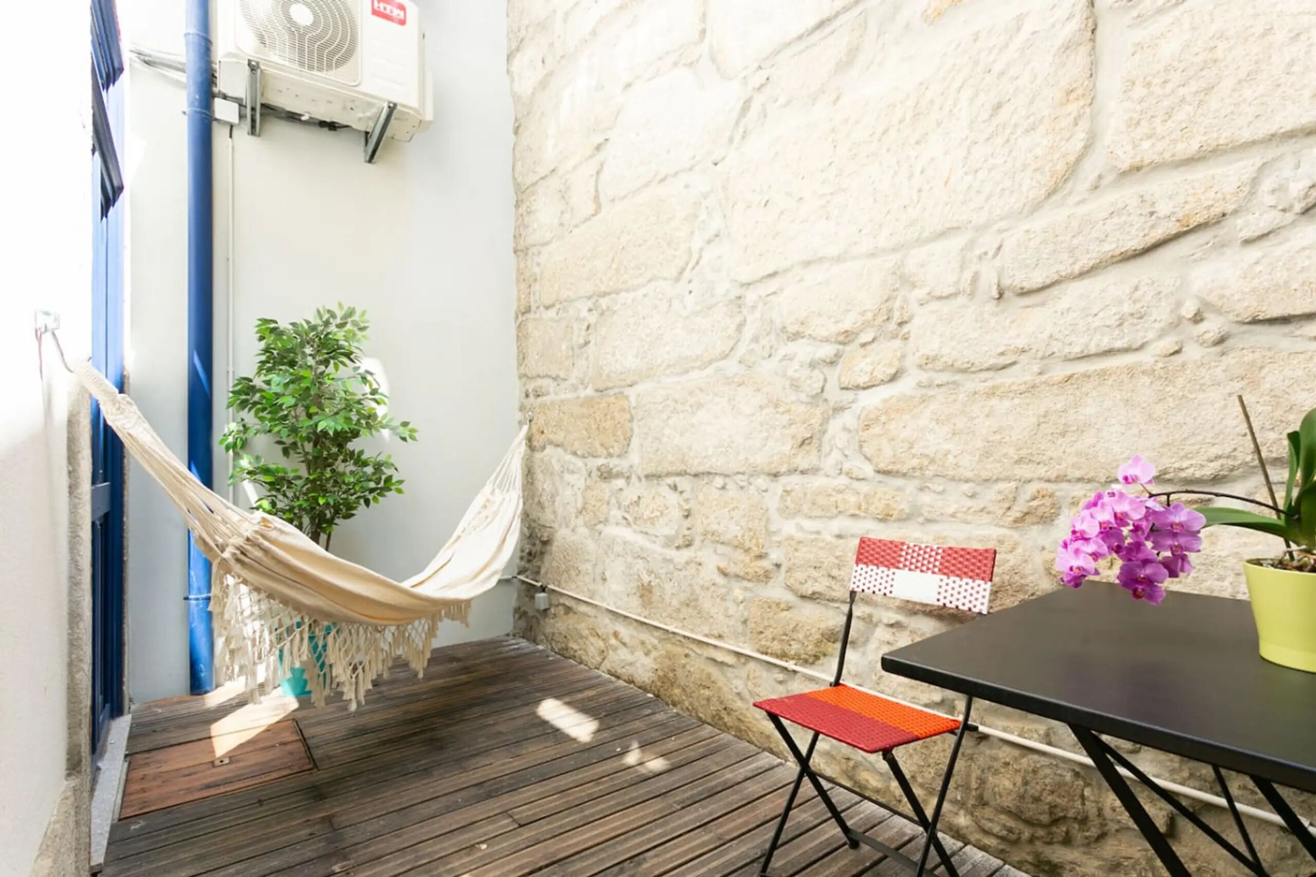 Eine Holzterrasse mit einer Hängematte, einem Blumentopf, einem kleinen Tisch mit einer Flagge und einer Steinsäule.
