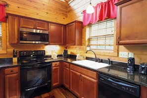 A kitchen with wooden cabinets, a stainless steel microwave, a black stove, a sink, and a window with blinds. - Gorgeous log cabin that sleeps 7 with hot tub, firepit, exclusive hiking trails! (Sevierville)