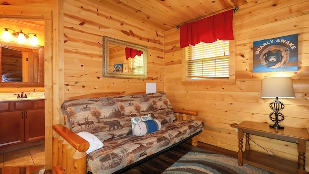 A cabin interior with a wooden interior, a sofa with a wildlife print, a small table with a lamp, a mirror, and a window with blinds. - Gorgeous log cabin that sleeps 7 with hot tub, firepit, exclusive hiking trails! (Sevierville)