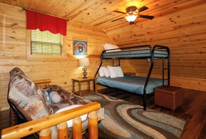 Ein gemütliches Blockhauszimmer mit Etagenbett, einem kleinen Tisch mit Lampe, einem roten Vorhang und einem an der Wand befestigten Deckenventilator.