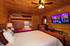 Habitación de cabaña de madera con literas, una cama individual, ventilador de techo, televisor mostrando la interfaz de un servicio de streaming y ventana con persianas.
