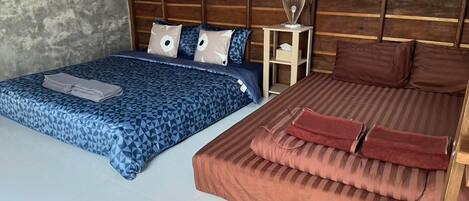 A bedroom with two beds, one with a patterned bedspread and the other with a striped bedspread. There is a small table with a lamp and a shelf with a decorative item.