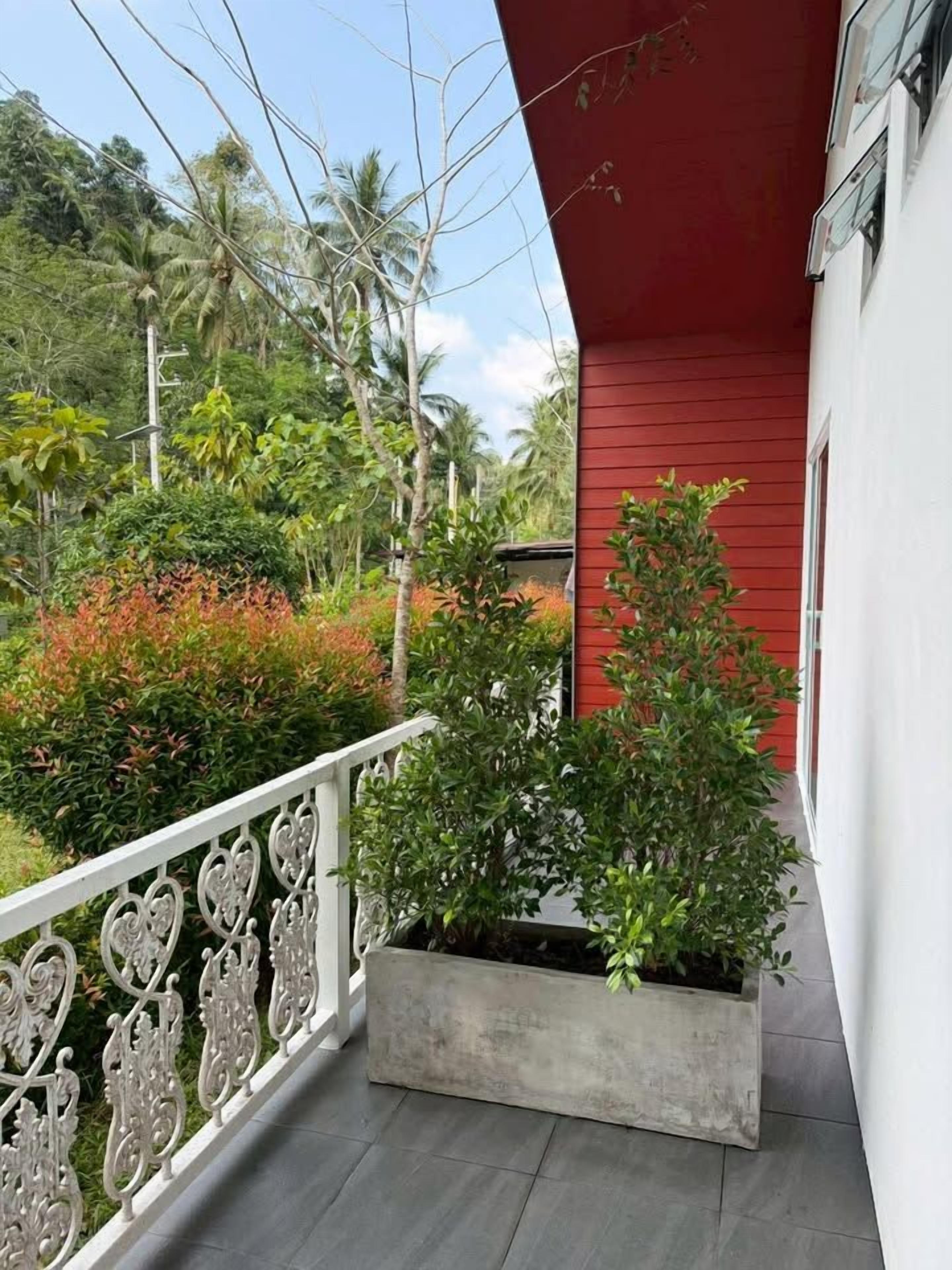 Un balcón con barandilla blanca, un edificio rojo y una jardinera con plantas verdes.