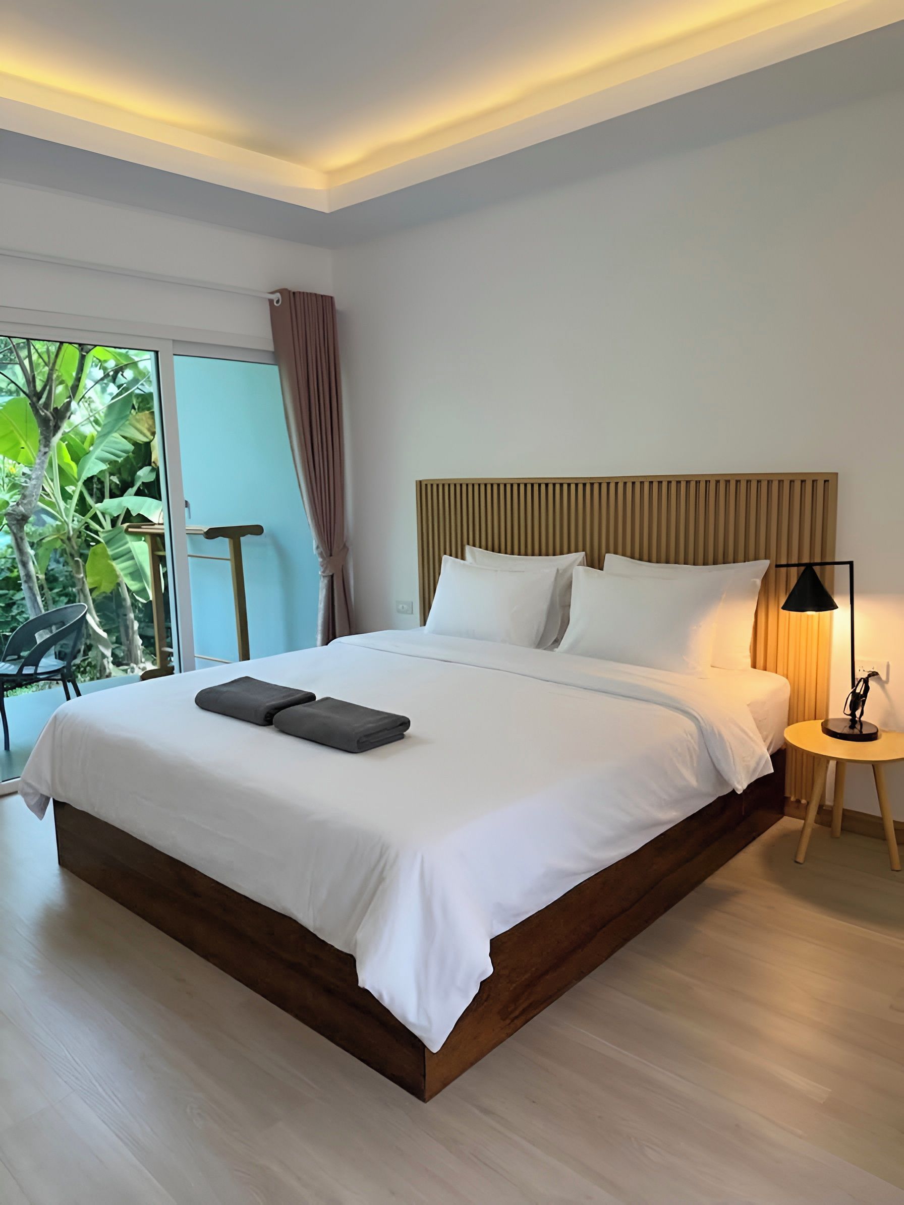 Une chambre d’hôtel moderne avec un grand lit, une table de chevet, une lampe et une vue sur la verdure environnante.