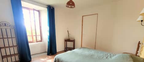 Een slaapkamer met een bed, houten vloeren, een raam met gordijnen, een deur en een hanglamp.
