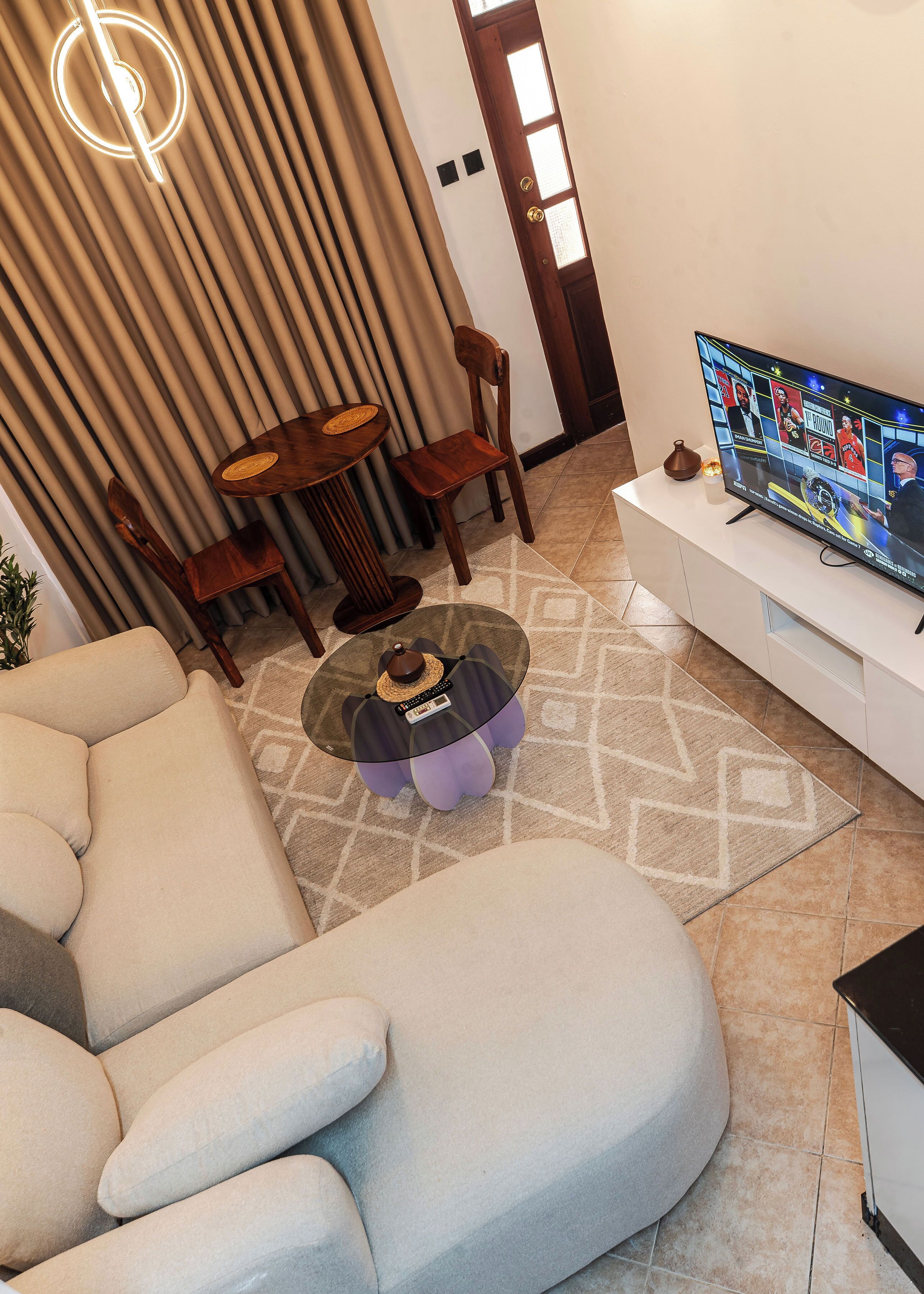 Una sala de estar moderna con un sofá modular, una mesa de centro con un control remoto, un televisor sobre un soporte blanco, y un comedor con una mesa y sillas de madera.