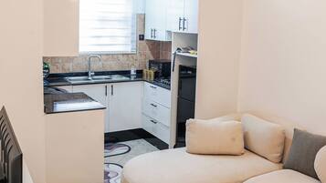 Una cocina moderna con armarios blancos, un microondas integrado y una estufa. En la sala de estar, hay un sofá color beige con cojines, y una mesa de centro de vidrio con un control remoto y un objeto decorativo.