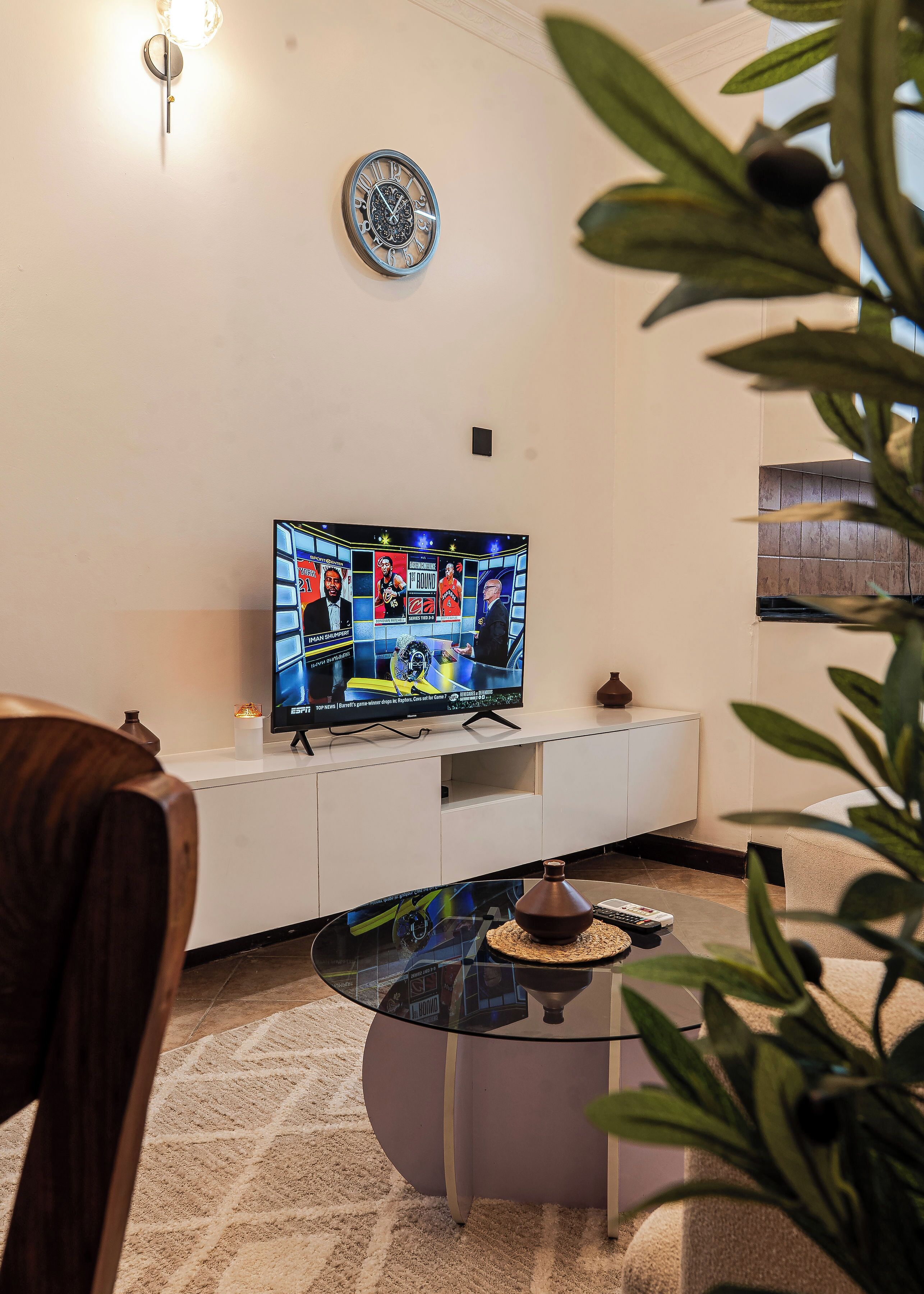 A living room with a flat-screen TV, a white entertainment unit, a glass coffee table, and a potted plant.