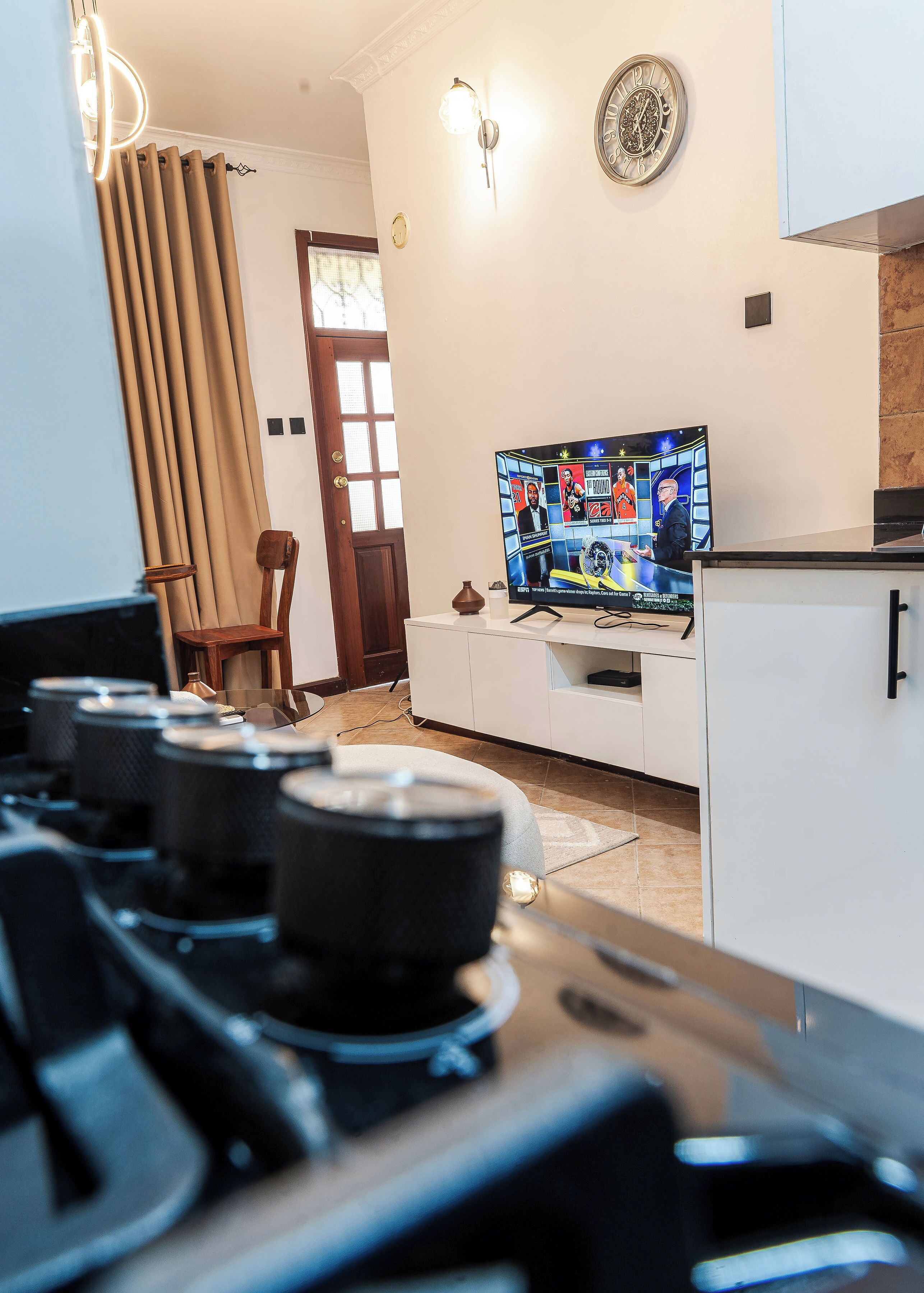 A modern kitchen with a gas stove, a television displaying sports content, and a dining area with a chair and a cabinet.
