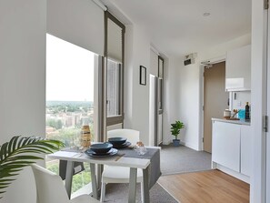 Eine moderne Küche mit weißen Schränken, ein Essbereich mit Tisch und Stühlen sowie ein Blick auf die Stadt durch große Fenster.