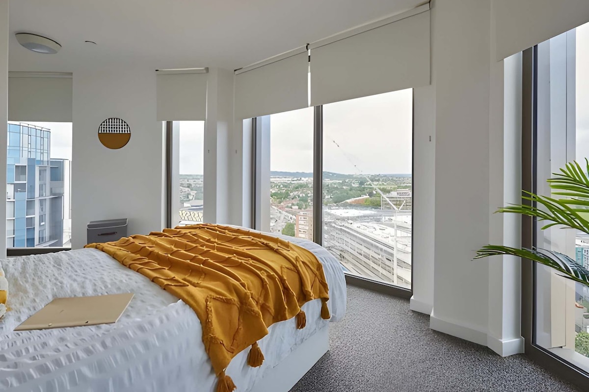 A modern hotel room with a large bed, a view of a cityscape, and a plant.