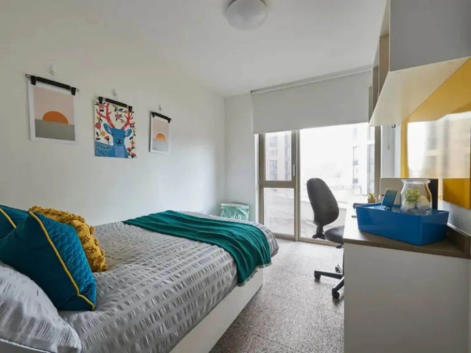 Ein Schlafzimmer mit einem Bett, einem Schreibtisch und einem Stuhl. Es gibt Fenster und Kunstwerke an der Wand.