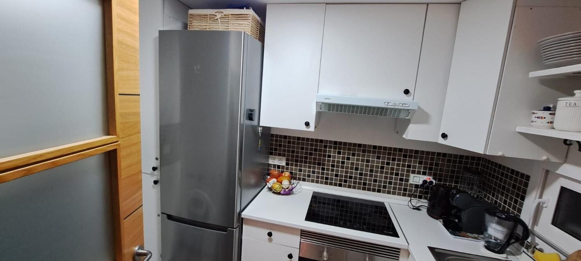 Eine kleine Küche mit Edelstahl-Kühlschrank, weißen Schränken, einer gefliesten Spritzschutzwand und einem Spülbecken inklusive Wasserkocher.