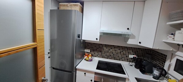 Una cocina compacta con refrigerador de acero inoxidable, armarios blancos, salpicadero de azulejos y un lavamanos con hervidor.