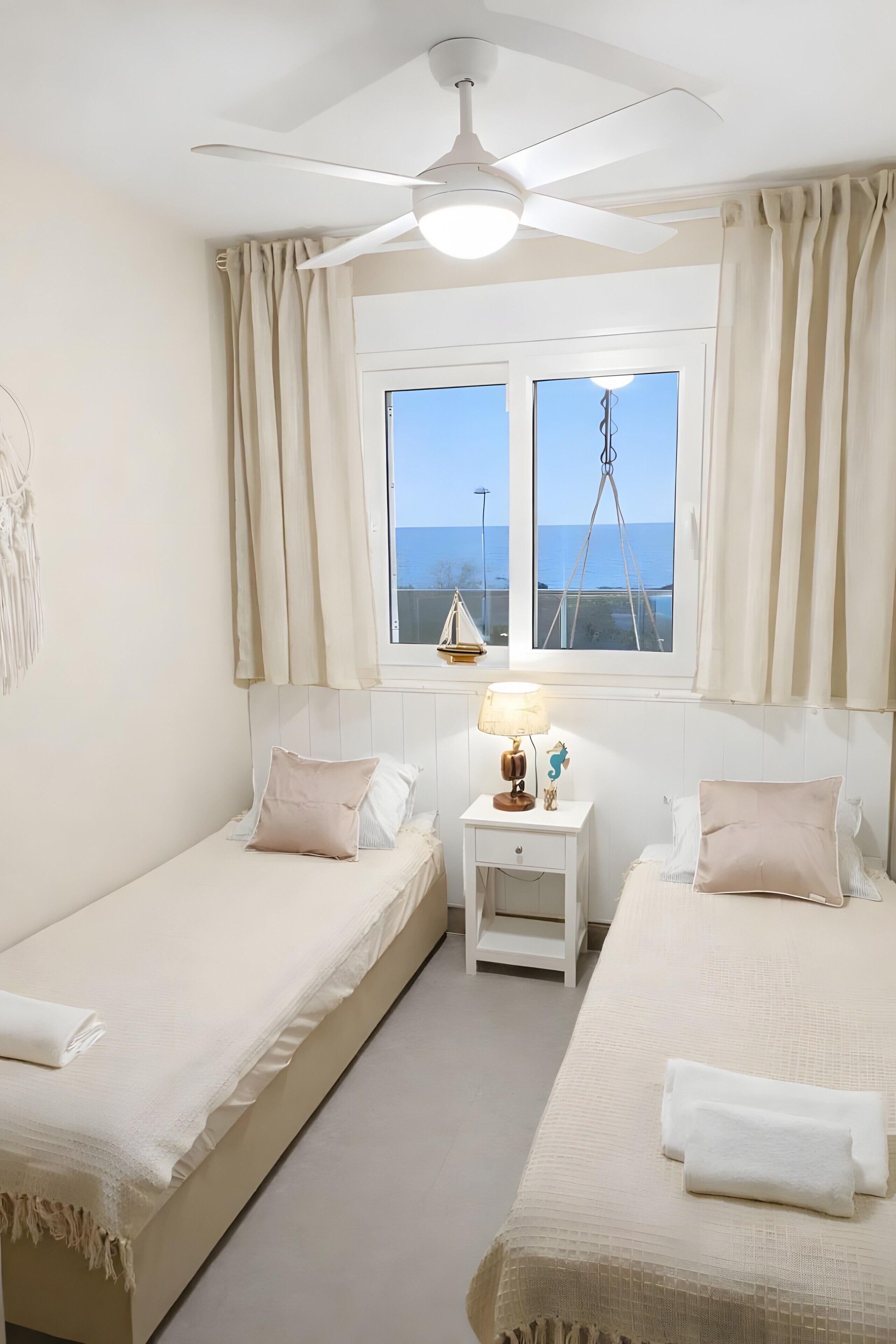 A bedroom with two beds, a ceiling fan, a window with curtains, and a nightstand with a lamp.
