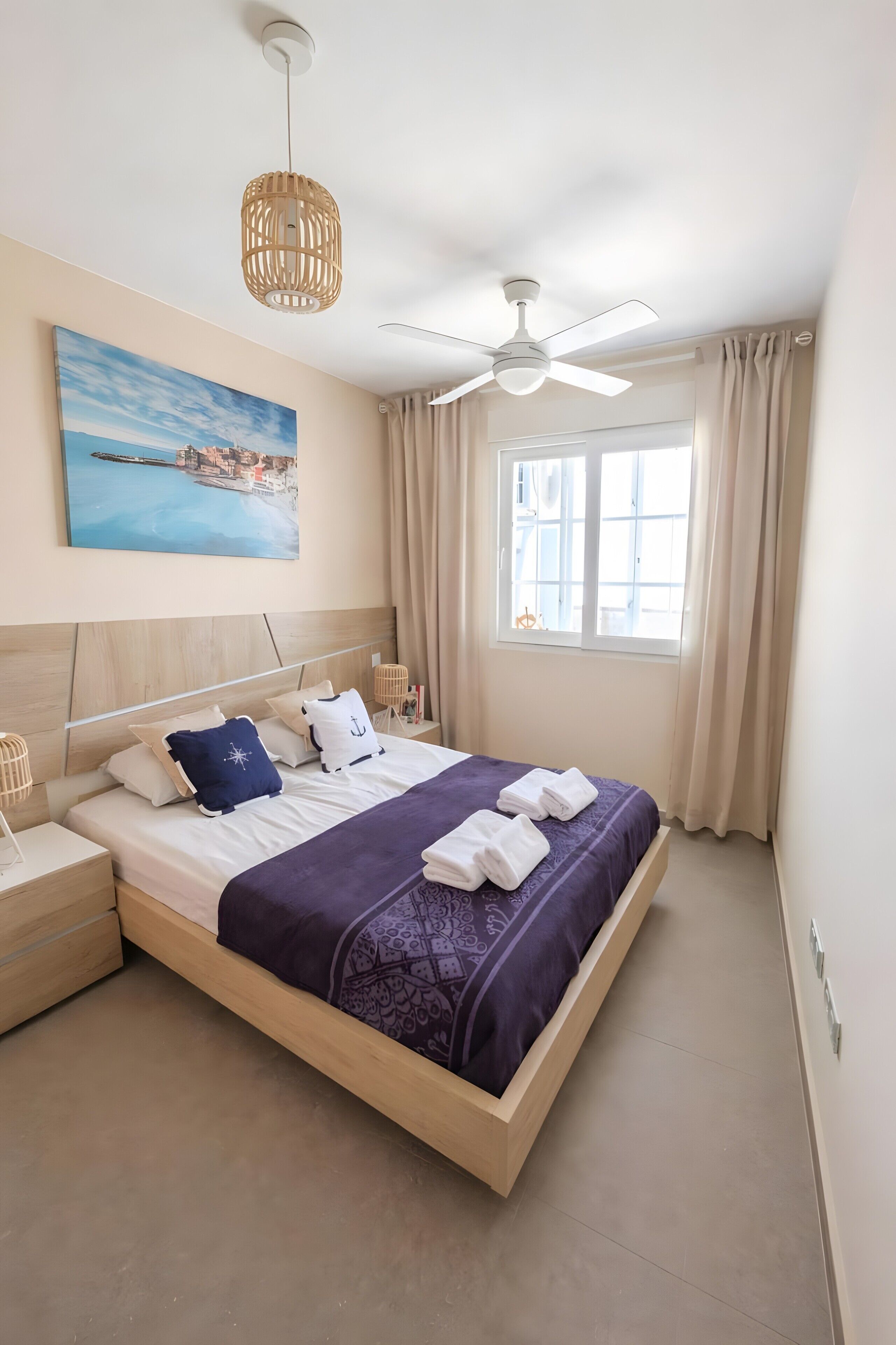 A neatly arranged hotel room with a bed, a nightstand, a lamp, a ceiling fan, a window with curtains, and a painting on the wall.