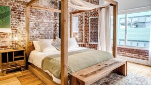 A four-poster bed with a wooden frame and white bedding, set against a brick wall with a large window. - Central City 2bed with Mountain Views Skywood Loft (Cape Town)