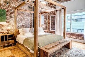 Ein Himmelbett mit Holzrahmen und weißer Bettwäsche, vor einer Ziegelwand mit einem großen Fenster.