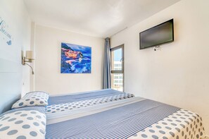 Ein Hotelzimmer mit Bett, Flachbildfernseher, einem Gemälde einer Küstenlandschaft, einem Fenster mit Vorhängen und einer Wandlampe.