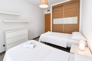 Et hotelværelse med to senge, en hvid kommode, et træskab og et natbord med en lampe.