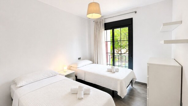 Quarto de hotel com duas camas, uma mesa de cabeceira com abajur, uma janela com cortinas e um guarda-roupa.