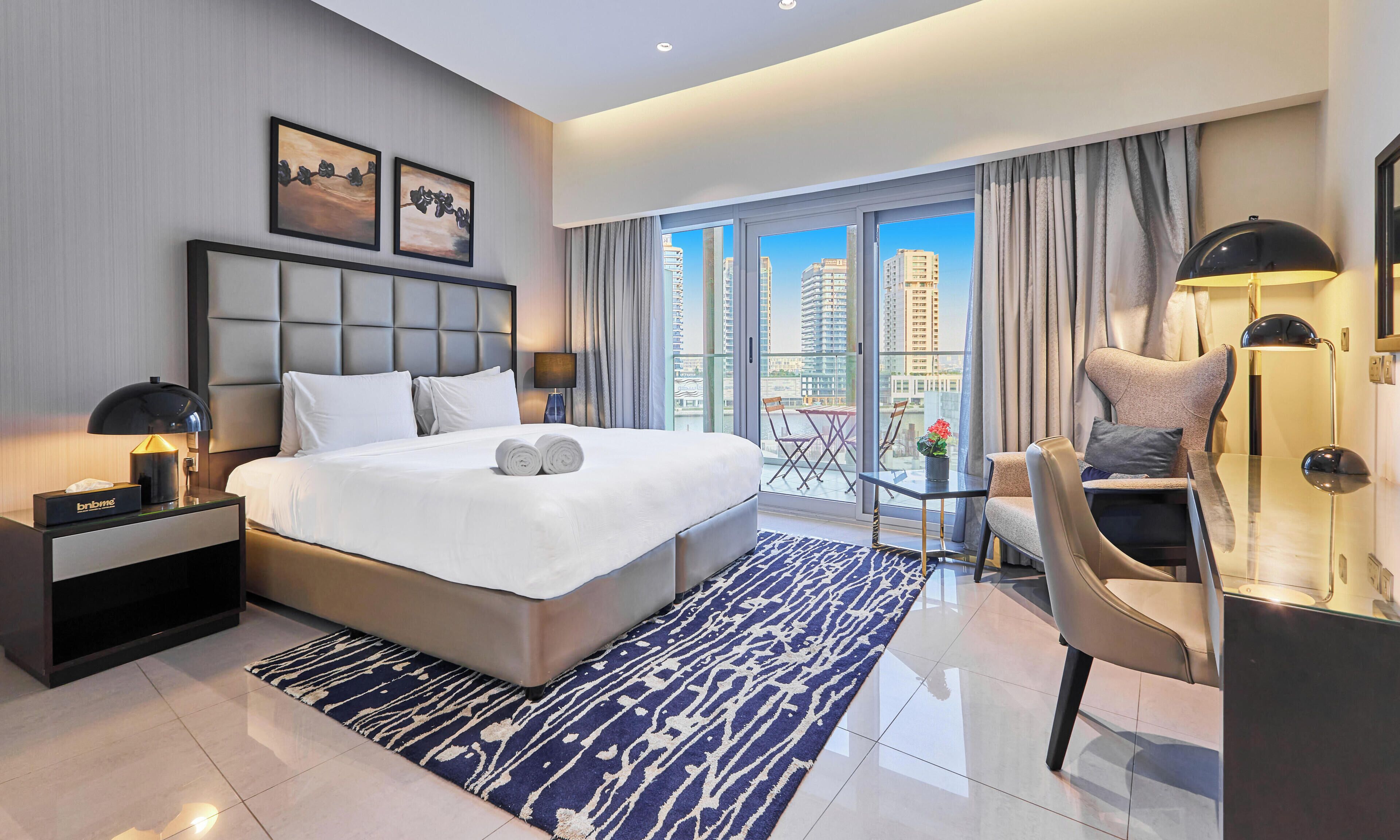 A hotel room with a large bed, a desk, a chair, a balcony with a view of buildings, and artwork on the wall.