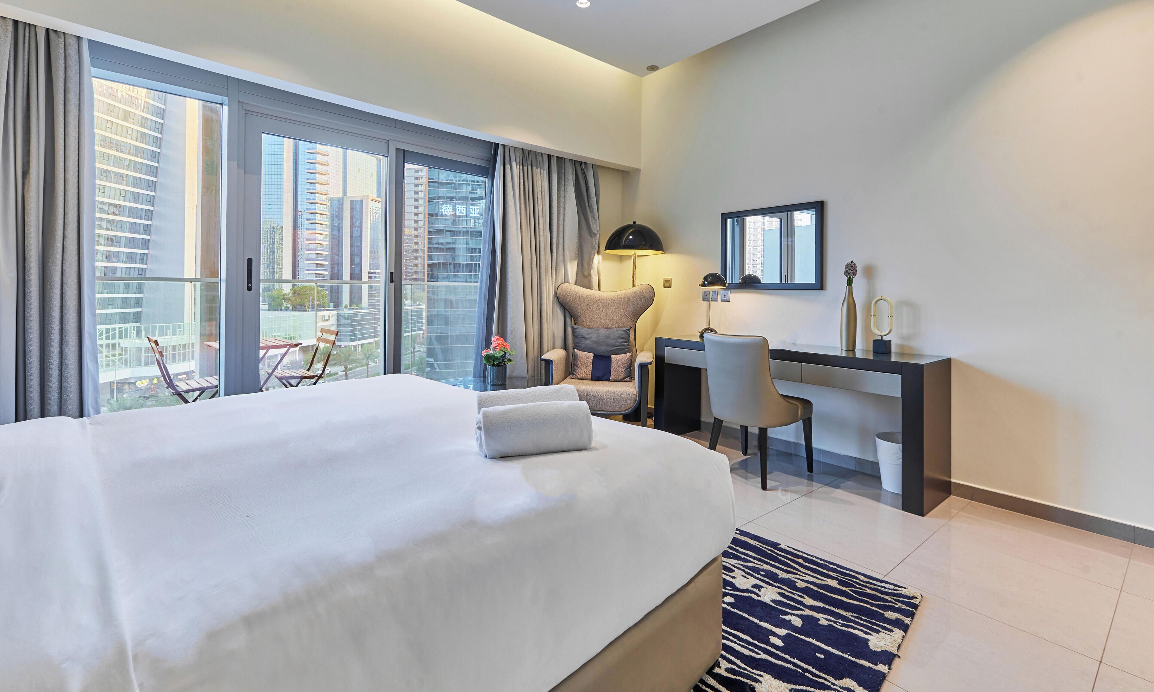 Une chambre d’hôtel équipée d’un lit, d’un bureau, d’une chaise, d’une lampe, d’un miroir et offrant une vue sur le paysage urbain grâce à de grandes fenêtres.