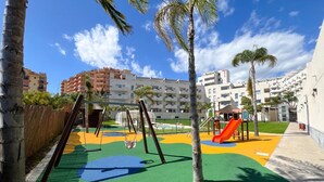 A playground with swings and slides, surrounded by palm trees and buildings. - Family apartment with sea views in Mijas Costa (Sitio de Calahonda)
