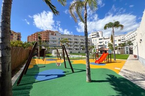 A playground with swings and slides, surrounded by palm trees and buildings. - Family apartment with sea views in Mijas Costa (Sitio de Calahonda)