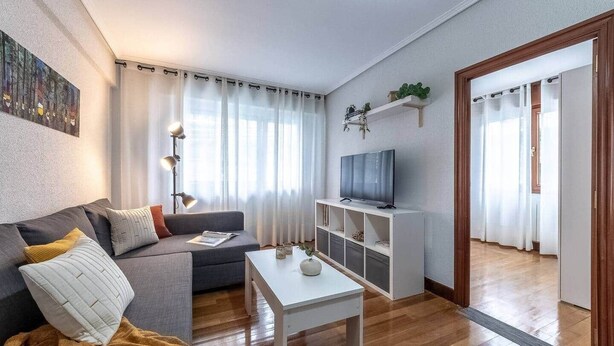 Un saló modern amb un sofà, una taula de centre, una televisió sobre un suport i una porta que dona a un altre espai.