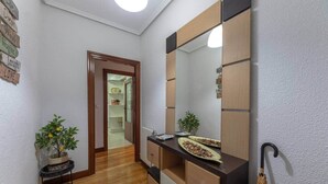 Un passadís amb terra de fusta, un mirall, una petita planta i una porta que dóna a una habitació amb una prestatgeria i un rellotge.