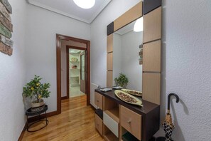 A hallway with a wooden floor, a mirror, a small plant, and a door leading to a room with a shelf and a clock. - POLITTE_Old Town vibes & parking (Bilbao)
